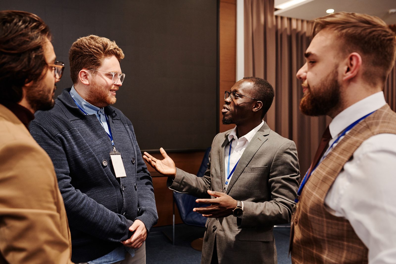 Four men in suits talk at an event; the man in the middle gestures with his hands.