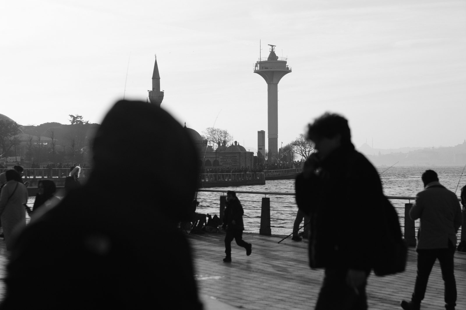 Black and white: silhouettes of people on a waterfront, with a lighthouse and a minaret behind.