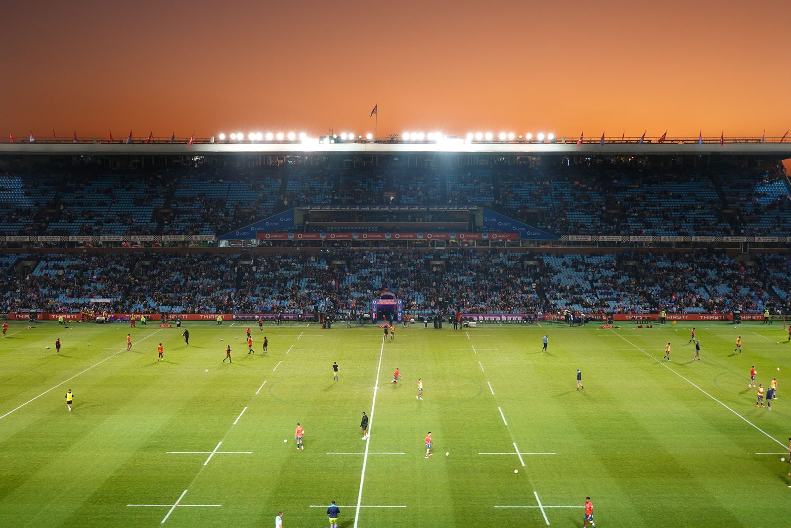 A rugby match in a large stadium under floodlights at sunset.