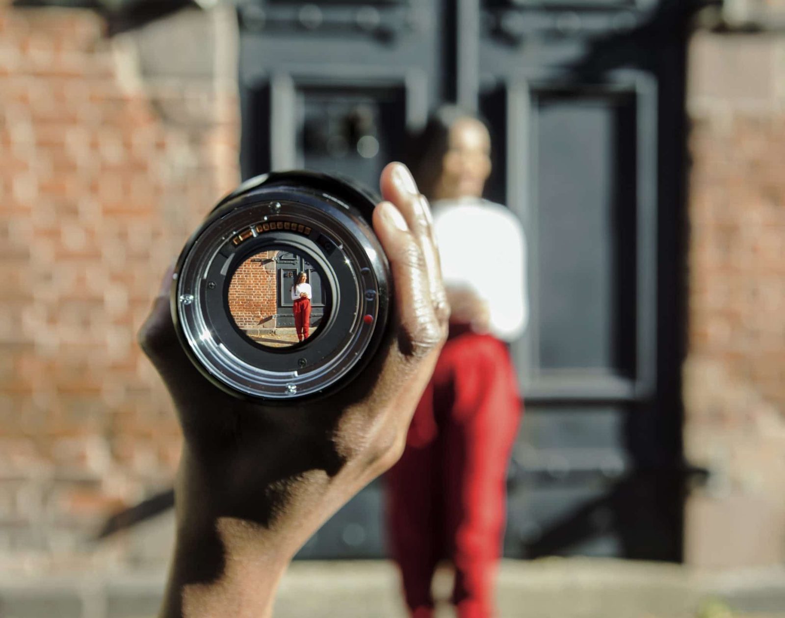 A hand holds a camera lens up to the camera; a person stands blurred in the background.