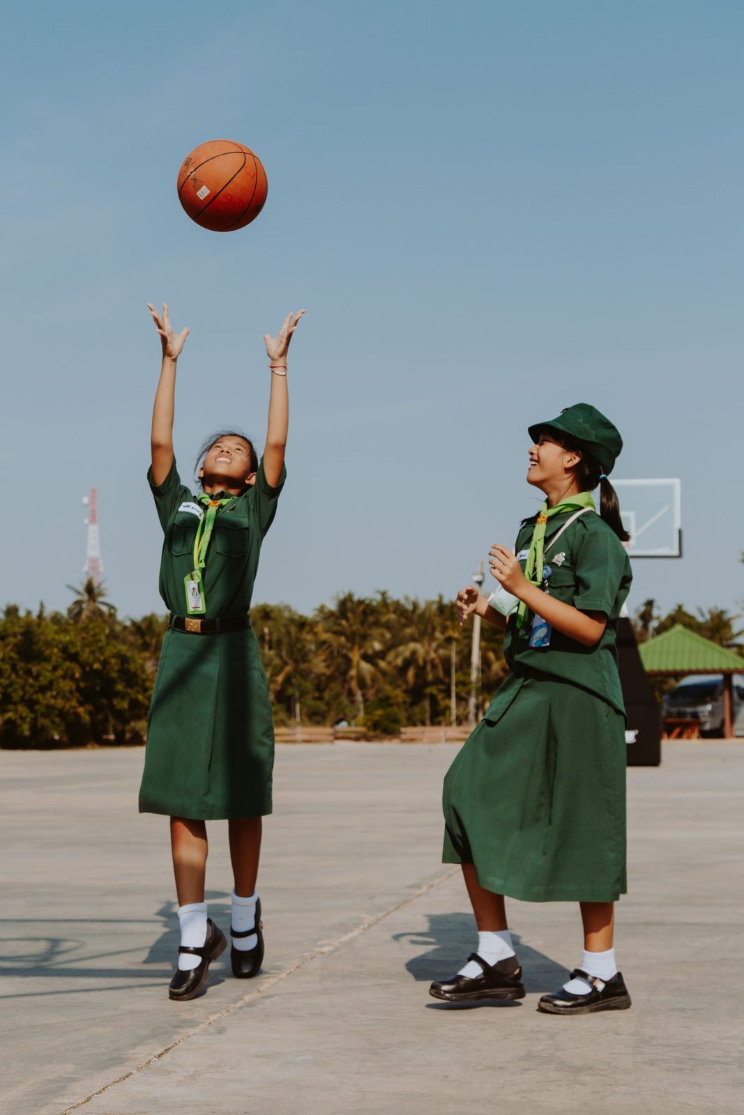 Two girls in green school uniforms play with a basketball; one throws it up.