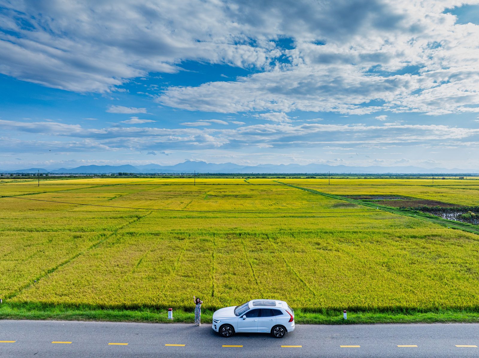 A small white car on a road along wide green rice fields under a cloudy sky.