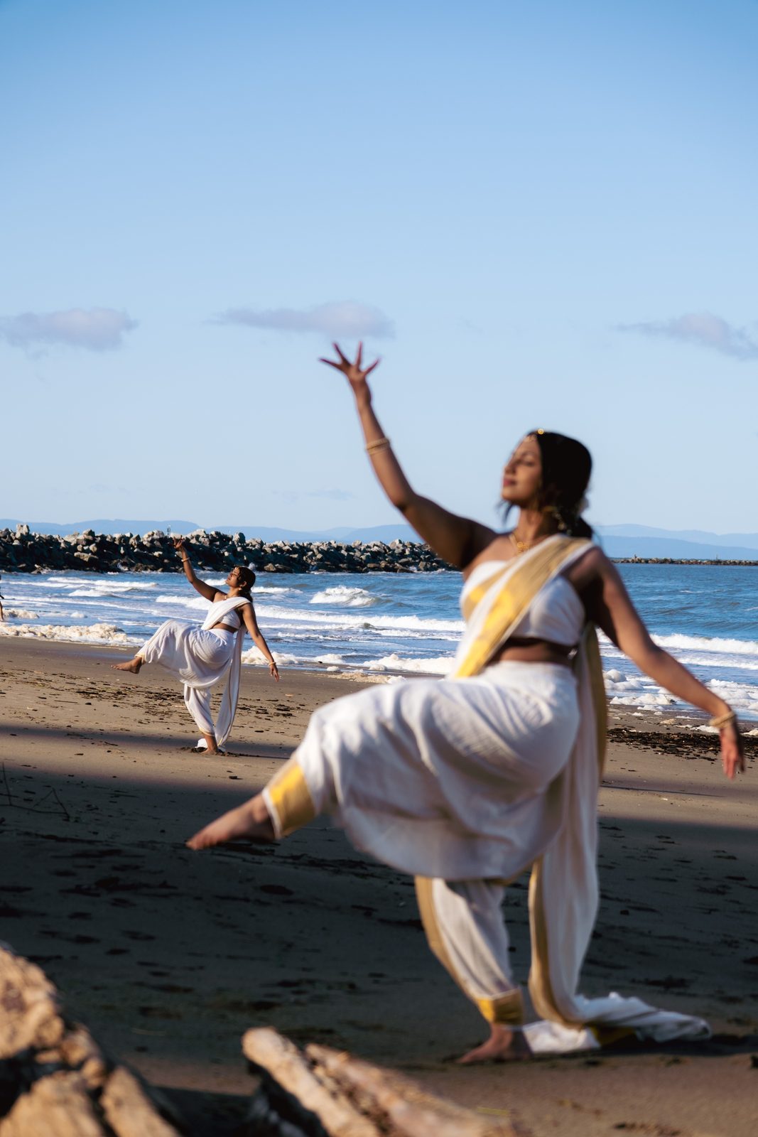 A woman in a white sari dances on a beach with one arm raised; a small child stands further back.