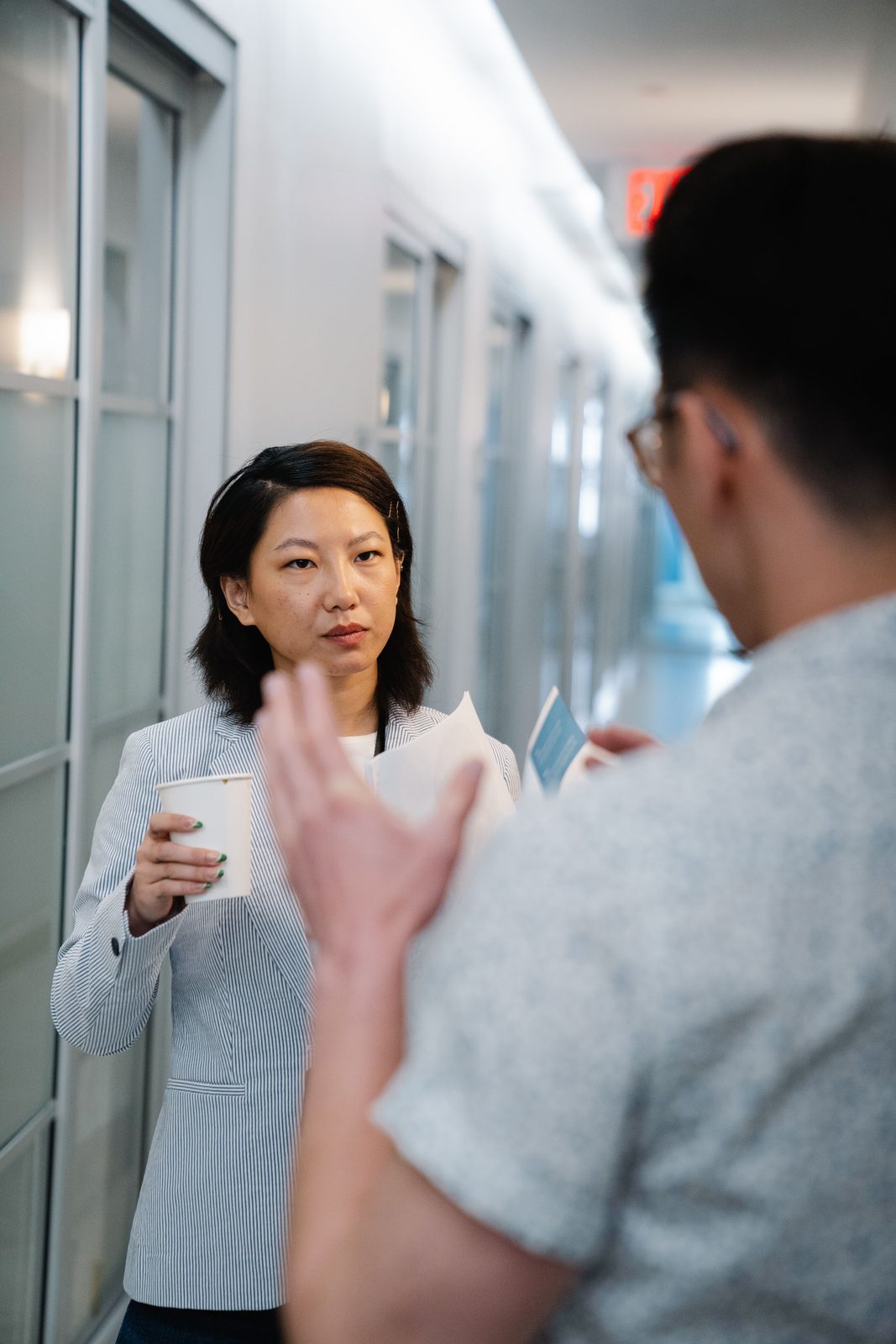 A woman holding a coffee cup listens to a man seen from behind, in a white corridor.