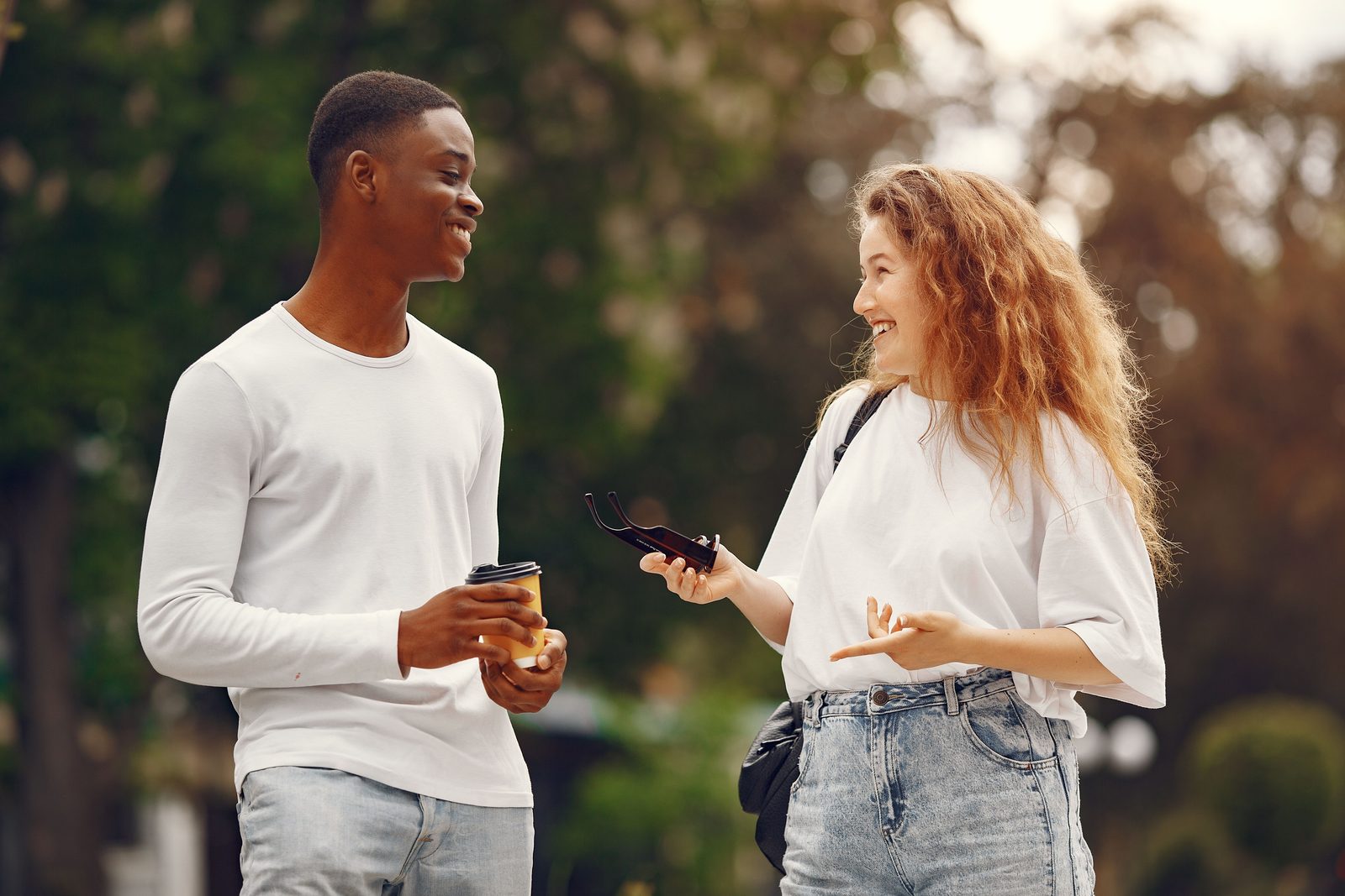 A young man and a woman with curly hair talk outdoors under trees; she holds a phone.